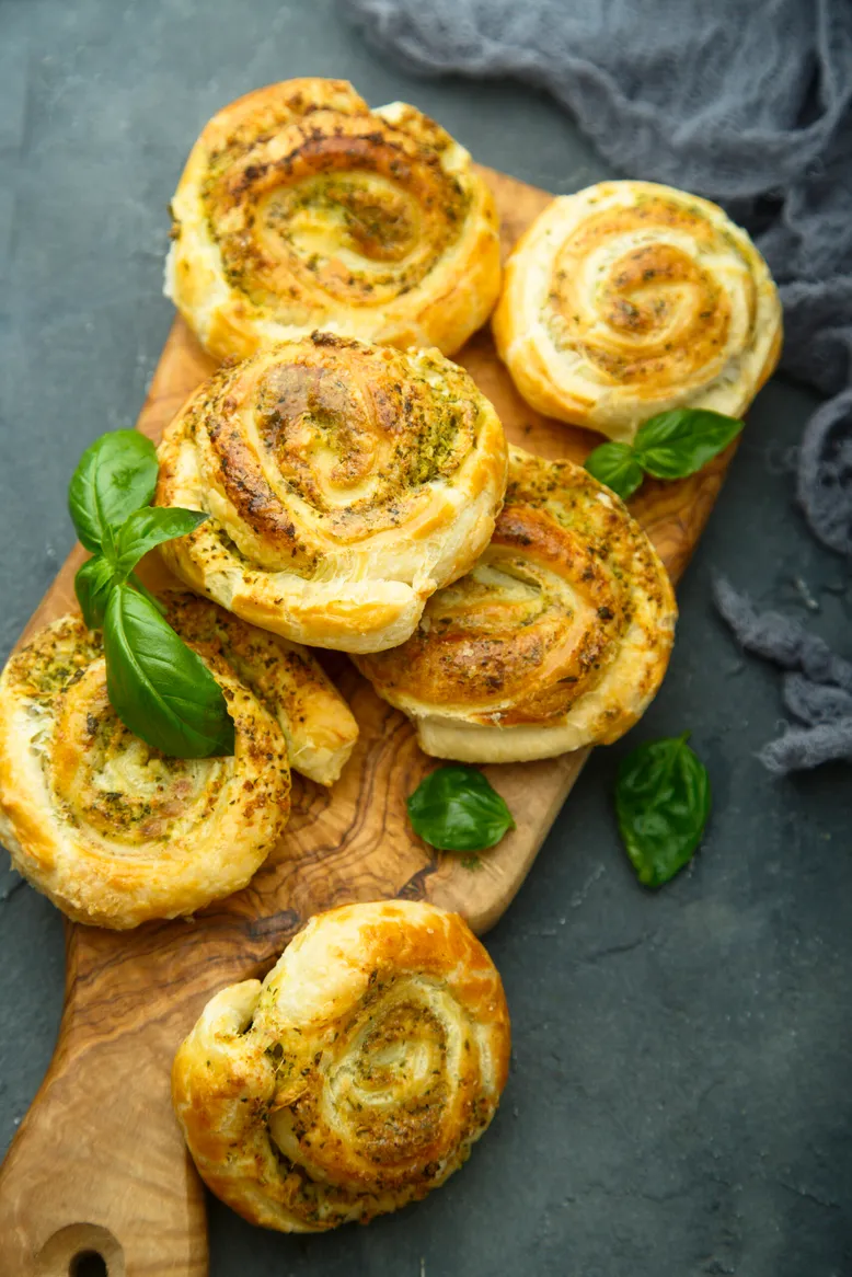 Escargots feuilletés au pesto, garnis de feuilles de basilic frais, sur une planche en bois.