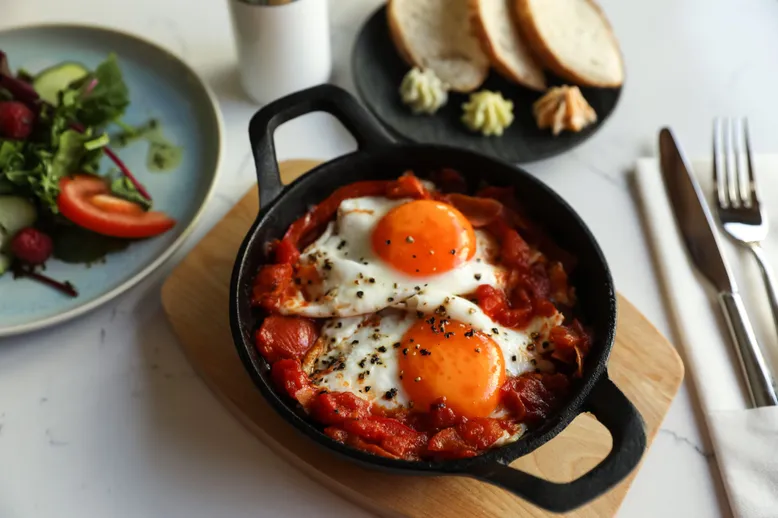 Shakshuka (œufs cuits dans une sauce tomate épicée) dans une poêle en fonte, avec salade et pain.