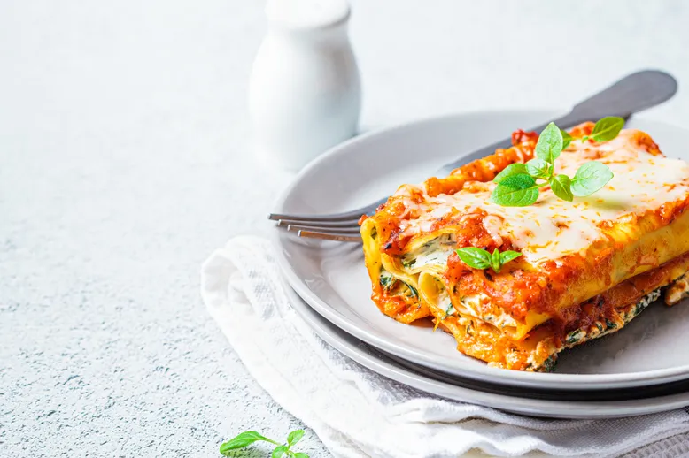 Part de lasagnes à la sauce tomate, fromage fondu et basilic frais, sur une assiette blanche.