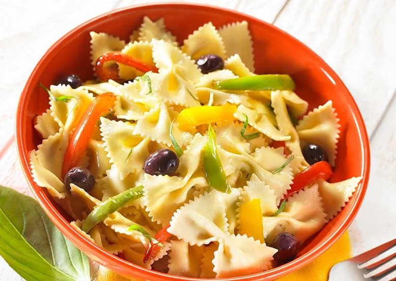 Bol rouge de salade de farfalle aux poivrons colorés, olives noires et basilic.