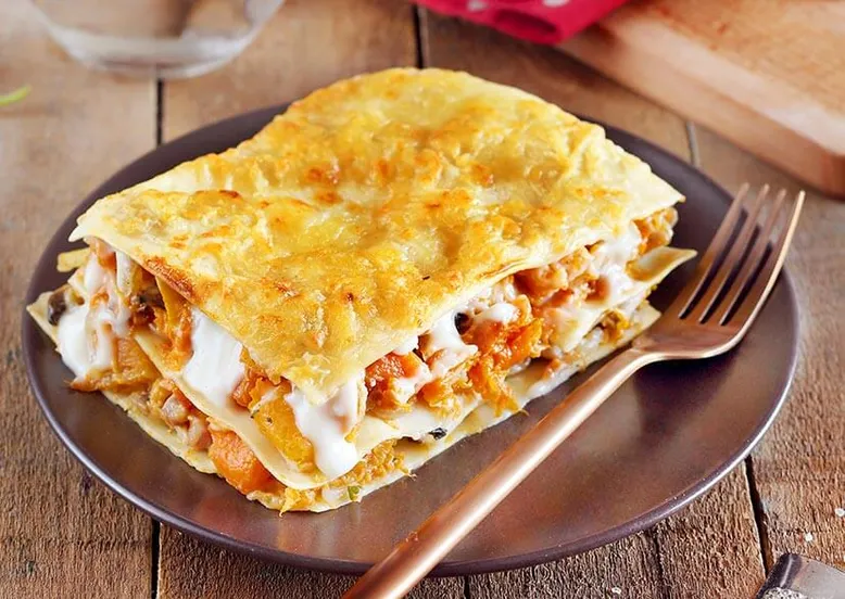 Part de lasagnes à la courge et sauce crémeuse, sur une assiette marron avec fourchette.