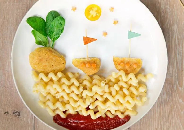 Assiette enfantine de pâtes en forme de vagues, nuggets de poulet en bateaux avec voiles d'épinards, soleil tomate.